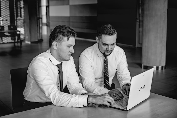 Dwóch mężczyzn w koszulach i krawatach pracuje wspólnie przy białym laptopie VAIO na jasnym stole w nowoczesnym wnętrzu z kolumnami, czarno-białe zdjęcie.