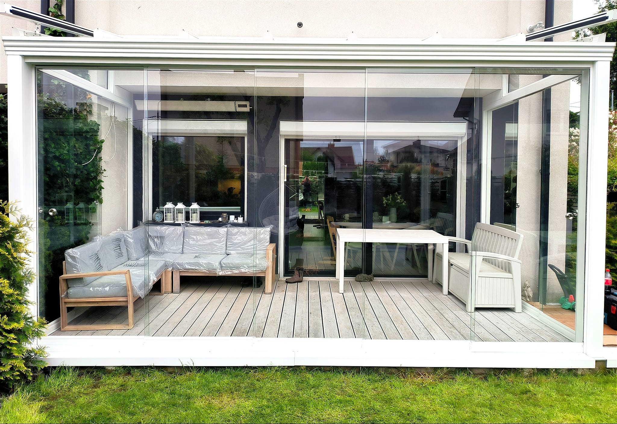 Zadaszony taras z drewnianą podłogą i szklanymi ścianami, meble ogrodowe częściowo zabezpieczone folią ochronną, widok na ogród.
