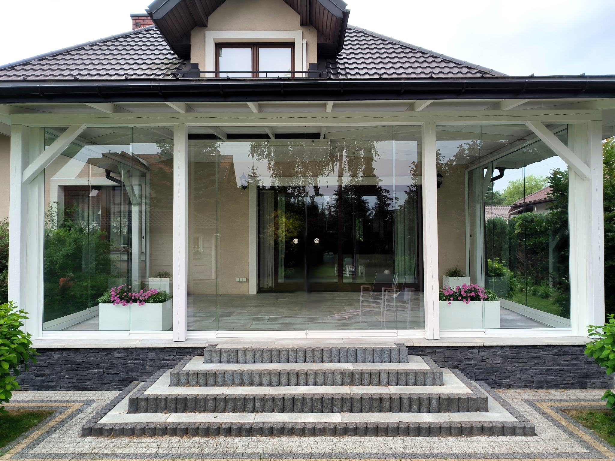 Zadaszony taras z przeszklonymi ścianami i schodami z kostki brukowej, donice z różowymi kwiatami, widok na ogród.