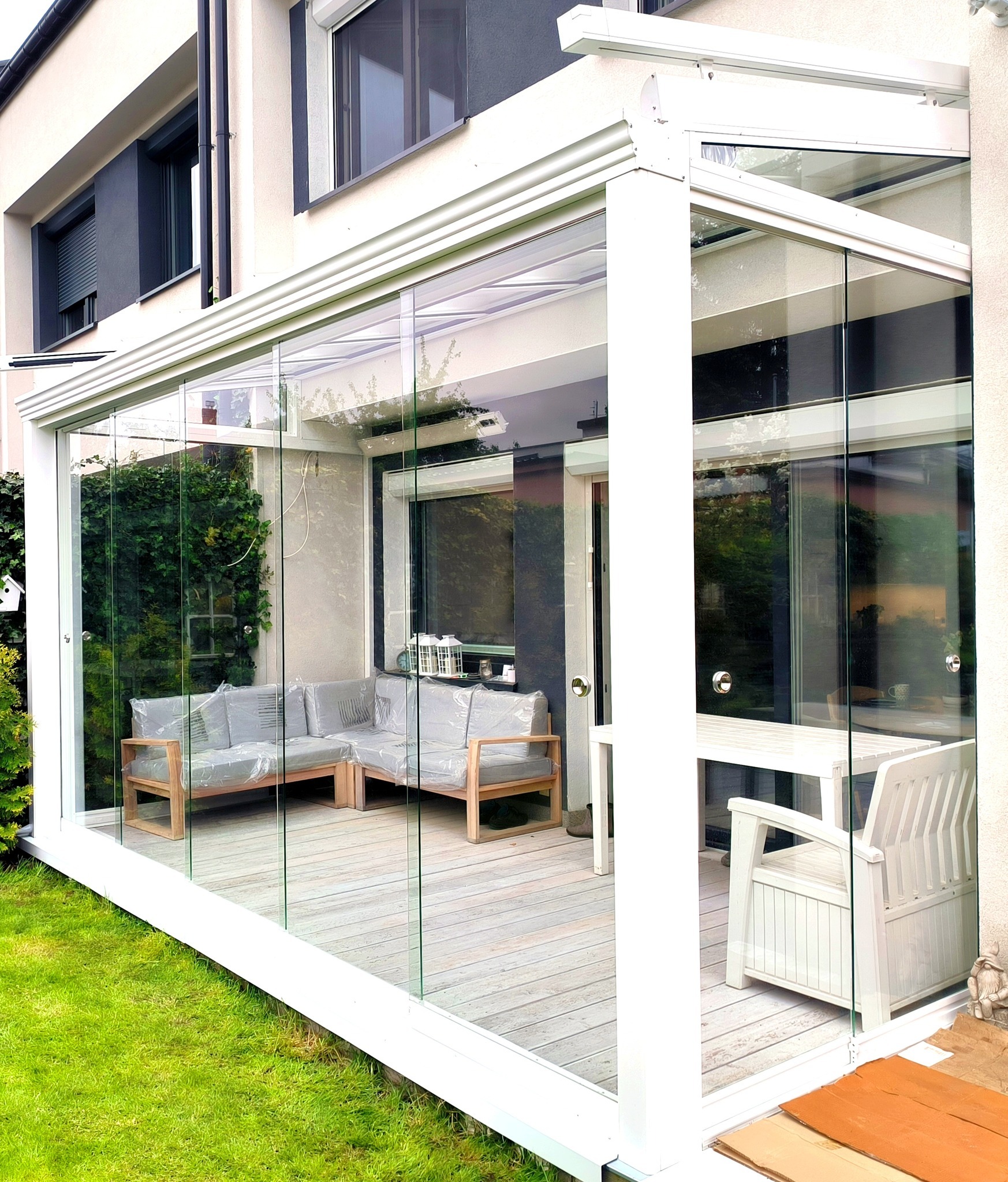 Szklana zabudowa tarasu z białymi ramami w Sulejówku, widoczne meble ogrodowe pod folią ochronną i drewniana podłoga tarasu.