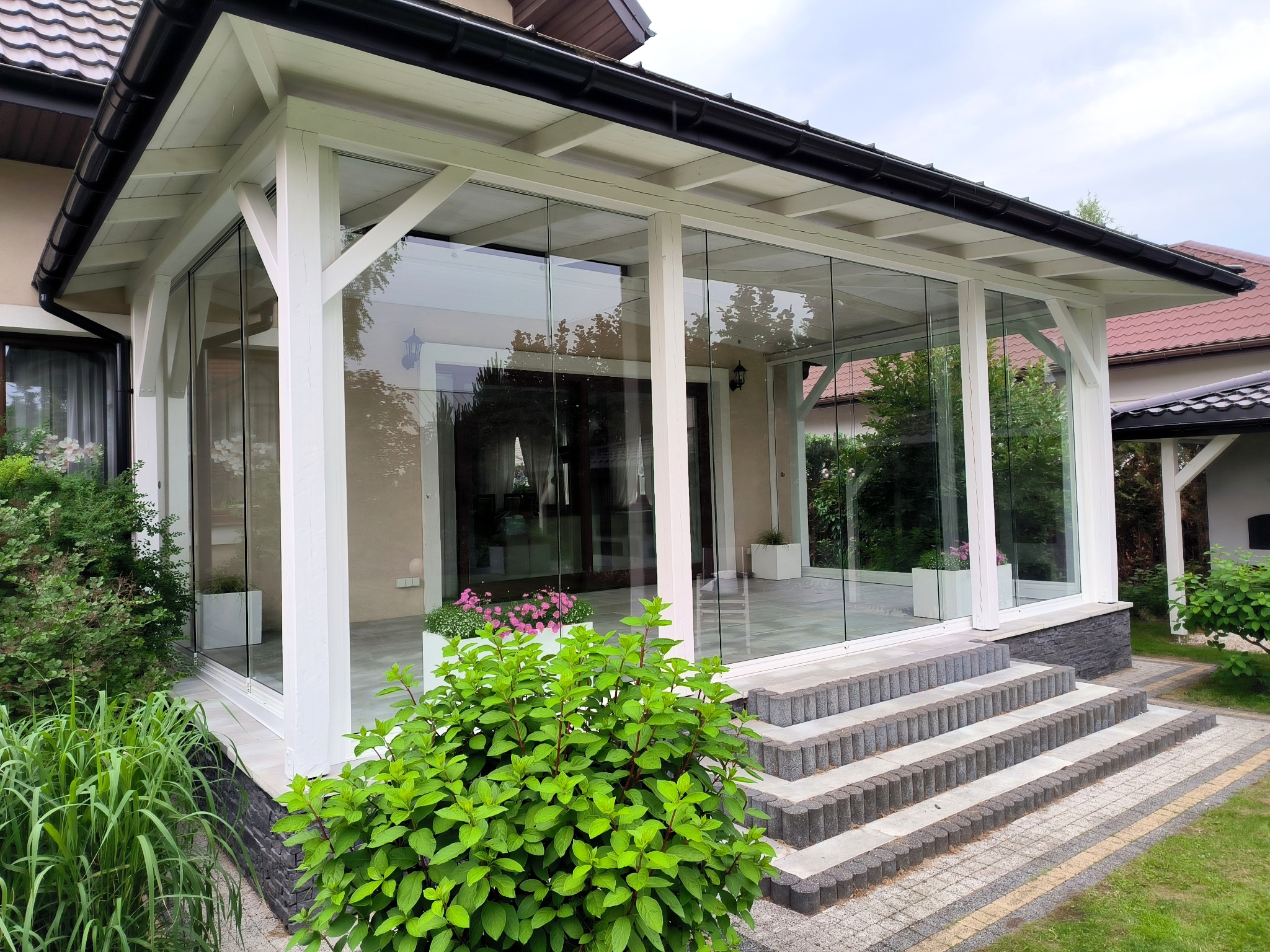 Szklana zabudowa tarasu z białymi, drewnianymi elementami konstrukcyjnymi, widoczne schody wejściowe i roślinność w ogrodzie.