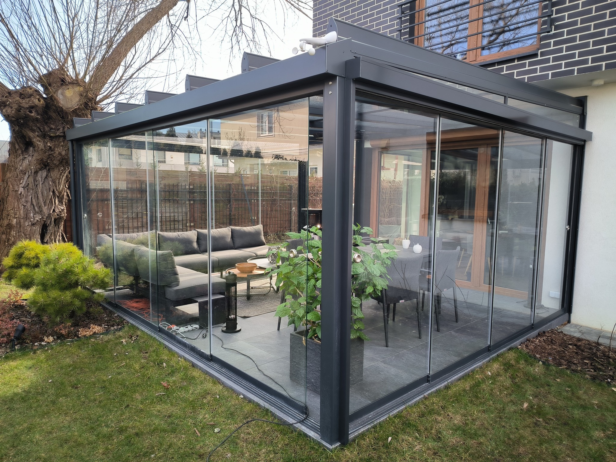 Zadaszony taras z przesuwnymi szklanymi ścianami, wyposażony w szarą sofę narożną i stół kawowy, z widokiem na ogród z drzewem.