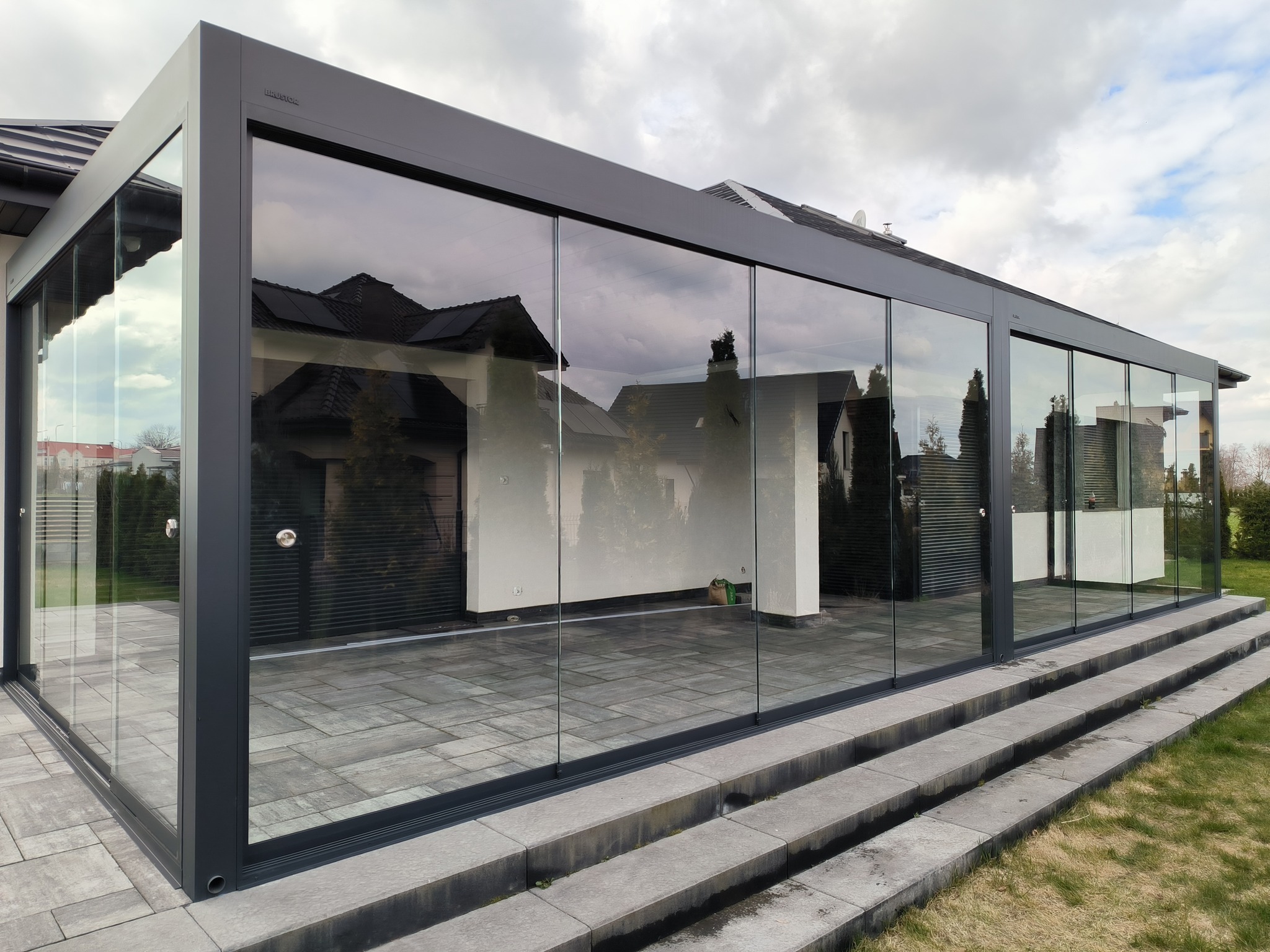 Nowoczesna, przeszklona konstrukcja z metalową ramą, prawdopodobnie z przesuwnymi drzwiami, na kamiennym tarasie, z widokiem na ogród i dom w tle, odbijającym się w szklanych panelach.