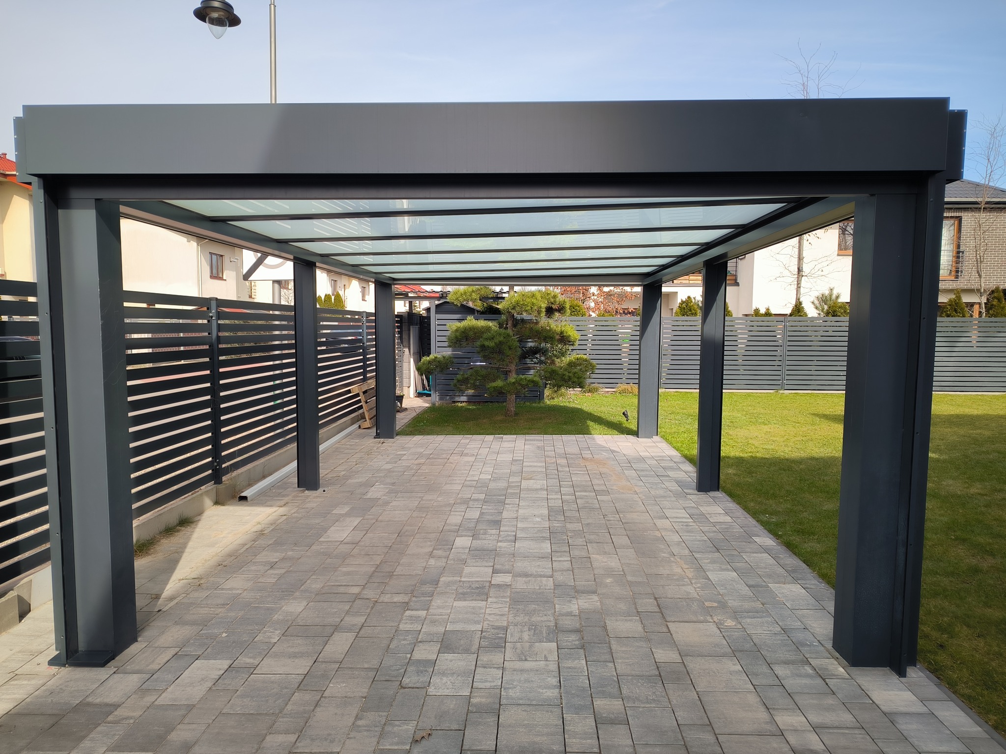 Nowoczesna, grafitowa konstrukcja z przeszklonym dachem, tworząca zadaszone miejsce parkingowe lub strefę relaksu na posesji z kostką brukową i zadbanym trawnikiem.