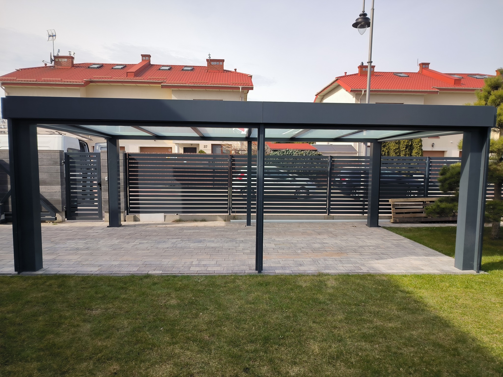 Nowoczesna, grafitowa konstrukcja z metalu z przeszklonym dachem, prawdopodobnie wiata garażowa, na tle domu z czerwoną dachówką i szarego ogrodzenia.