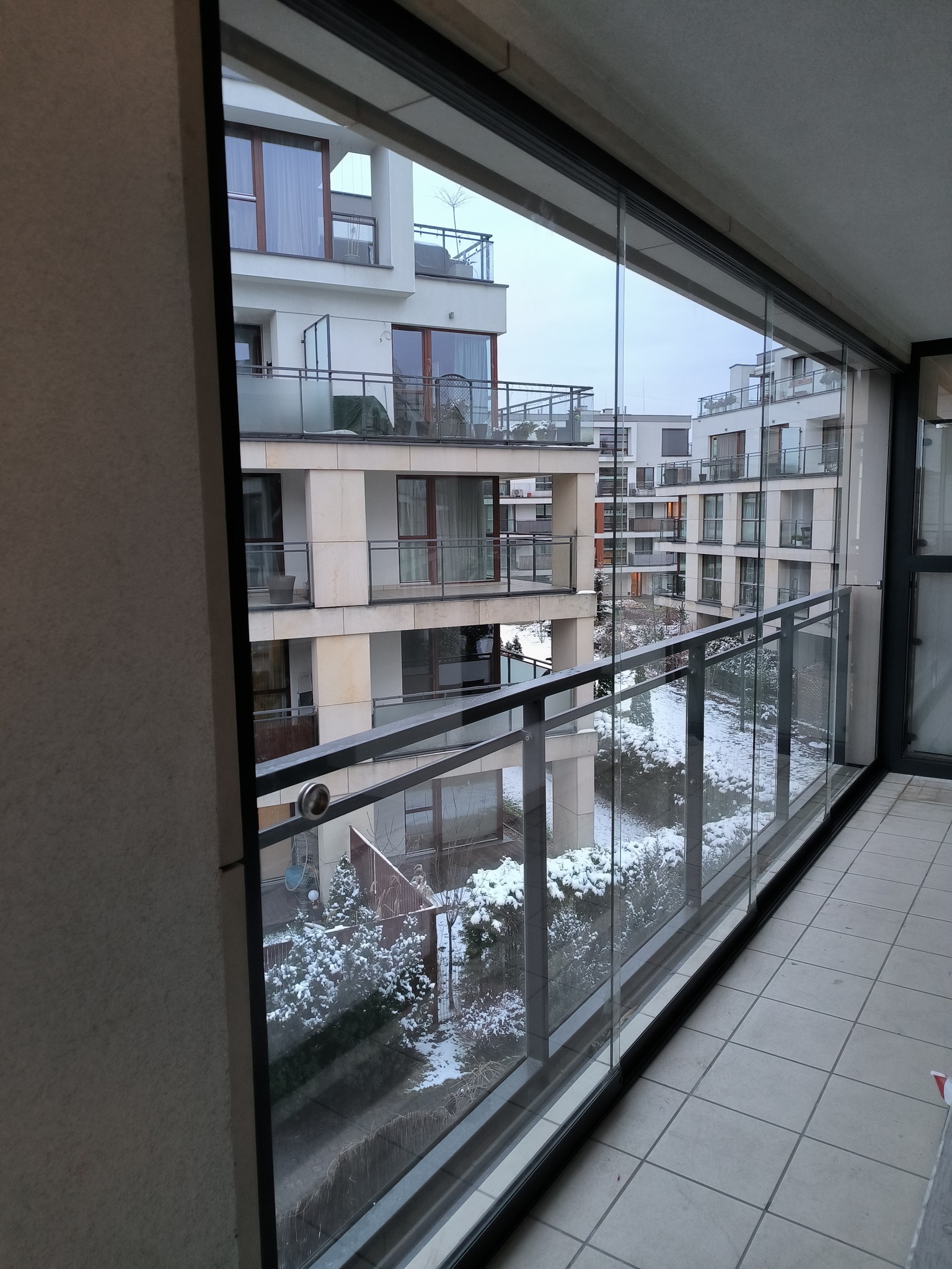 Zimowy widok z zadaszonego balkonu na osiedle apartamentowców, śnieg pokrywa drzewa i krzewy, widoczne szklane panele balkonowe i metalowa balustrada.