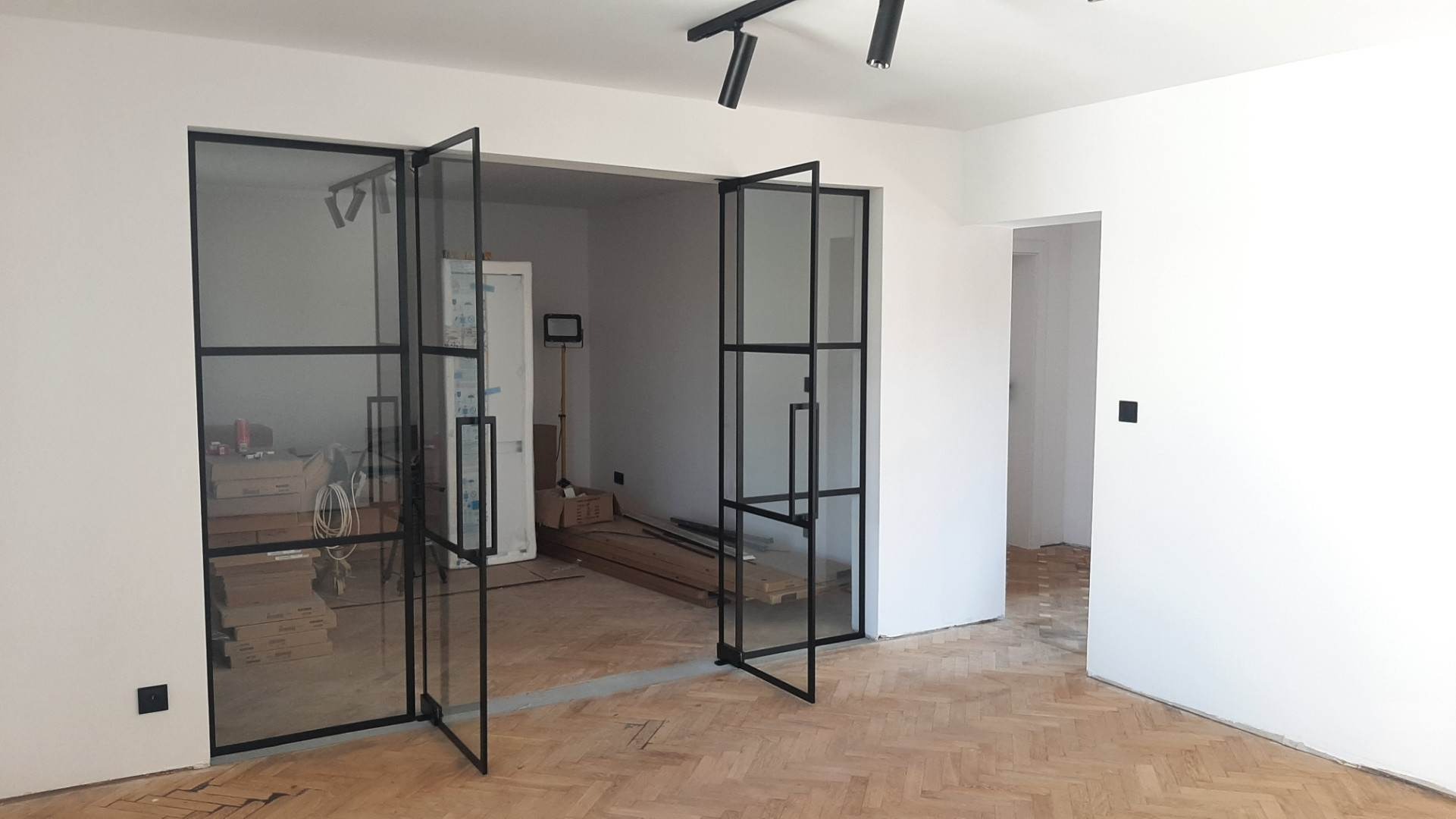 Wnętrze z podłogą w jodełkę i przeszklonymi, składanymi drzwiami w czarnych ramach, widoczne materiały budowlane w tle.