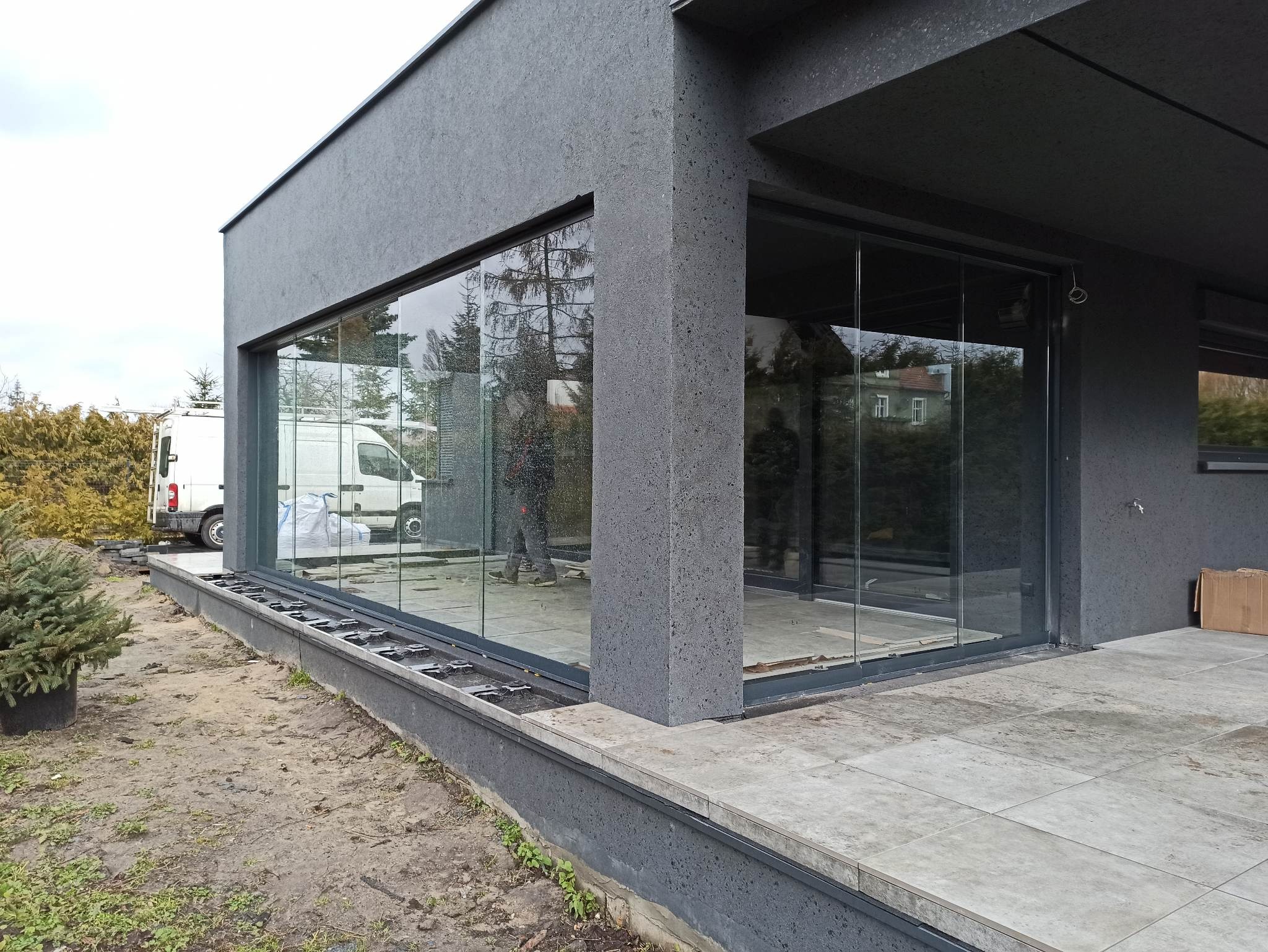 Montaż szklanej zabudowy tarasu w nowoczesnym domu z szarą elewacją, widoczny bus dostawczy i elementy konstrukcyjne tarasu kompozytowego.