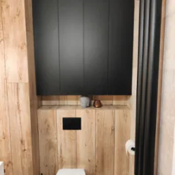 Nowoczesna toaleta z drewnopodobnymi płytkami, czarnymi szafkami w zabudowie i grzejnikiem drabinkowym w tym samym kolorze. Na półce dekoracje: głowa Buddy i świecznik.