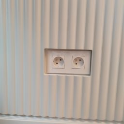 HOHO PROJEKT Adam Śleszyński - Biała, ryflowana ściana z dwoma gniazdkami elektrycznymi w białej ramce. Widoczna listwa przypodłogowa i fragment podłogi. Minimalistyczny design wnętrza.