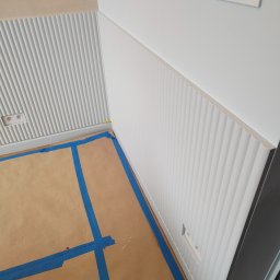 HOHO PROJEKT Adam Śleszyński - Montaż paneli ściennych z frezowaniem pionowym, zabezpieczona podłoga kartonem i taśmą malarską. Widoczne gniazdka elektryczne i włącznik światła.