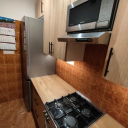 HOHO PROJEKT Adam Śleszyński - Nowoczesna kuchnia z drewnianymi frontami, srebrną lodówką i kuchenką gazową. Uwagę przykuwa brązowa glazura na ścianie i kalendarz na lodówce.