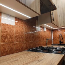 HOHO PROJEKT Adam Śleszyński - Nowoczesna kuchnia z drewnianym blatem, brązowymi płytkami i oświetleniem LED pod szafkami. Widoczna płyta gazowa i czarna bateria. Ujęcie z perspektywy blatu.