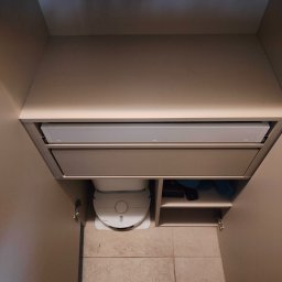 HOHO PROJEKT Adam Śleszyński - Wnętrze szafy wnękowej z szufladami i półkami, na podłodze robot sprzątający. Widoczne zawiasy drzwi i fragment buta osoby robiącej zdjęcie.