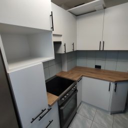 HOHO PROJEKT Adam Śleszyński - Nowoczesna kuchnia z białymi szafkami i drewnianym blatem. Czarne, podłużne uchwyty dodają elegancji. Widoczna płyta grzewcza i piekarnik. Jasne płytki na podłodze i ścianie.