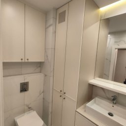 HOHO PROJEKT Adam Śleszyński - Jasna łazienka z zabudową meblową na wymiar: szafki wiszące, słupek z lamelami, umywalka z szufladami. Marmurowe płytki na ścianach, podświetlane lustro.