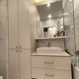 HOHO PROJEKT Adam Śleszyński - Jasna łazienka z szafką pod umywalką i wysoką szafą w zabudowie. Lustro z oświetleniem LED. Widoczne marmurowe płytki i fragment kabiny prysznicowej.