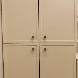 HOHO PROJEKT Adam Śleszyński - Zamknięte, kremowe fronty szafy z czterema drzwiczkami i okrągłymi, metalowymi uchwytami. Minimalistyczny design, gładkie powierzchnie, subtelne oświetlenie.