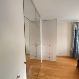 Biała szafa wnękowa z lustrzanymi drzwiami, odbijająca wnętrze pokoju z parkietem i zasłonami. Minimalistyczny design z okrągłymi uchwytami.