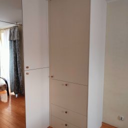 Biała szafa wnękowa z lustrzanymi drzwiami i drewnianymi uchwytami, widoczna w rogu pokoju. Minimalistyczny design, jasne wnętrze odbijające się w lustrze.