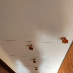 Biała szafa z drewnianymi uchwytami w mieszkaniu w Warszawie. Ujęcie z dołu, widoczne drewniane panele podłogowe i fragment drabiny.