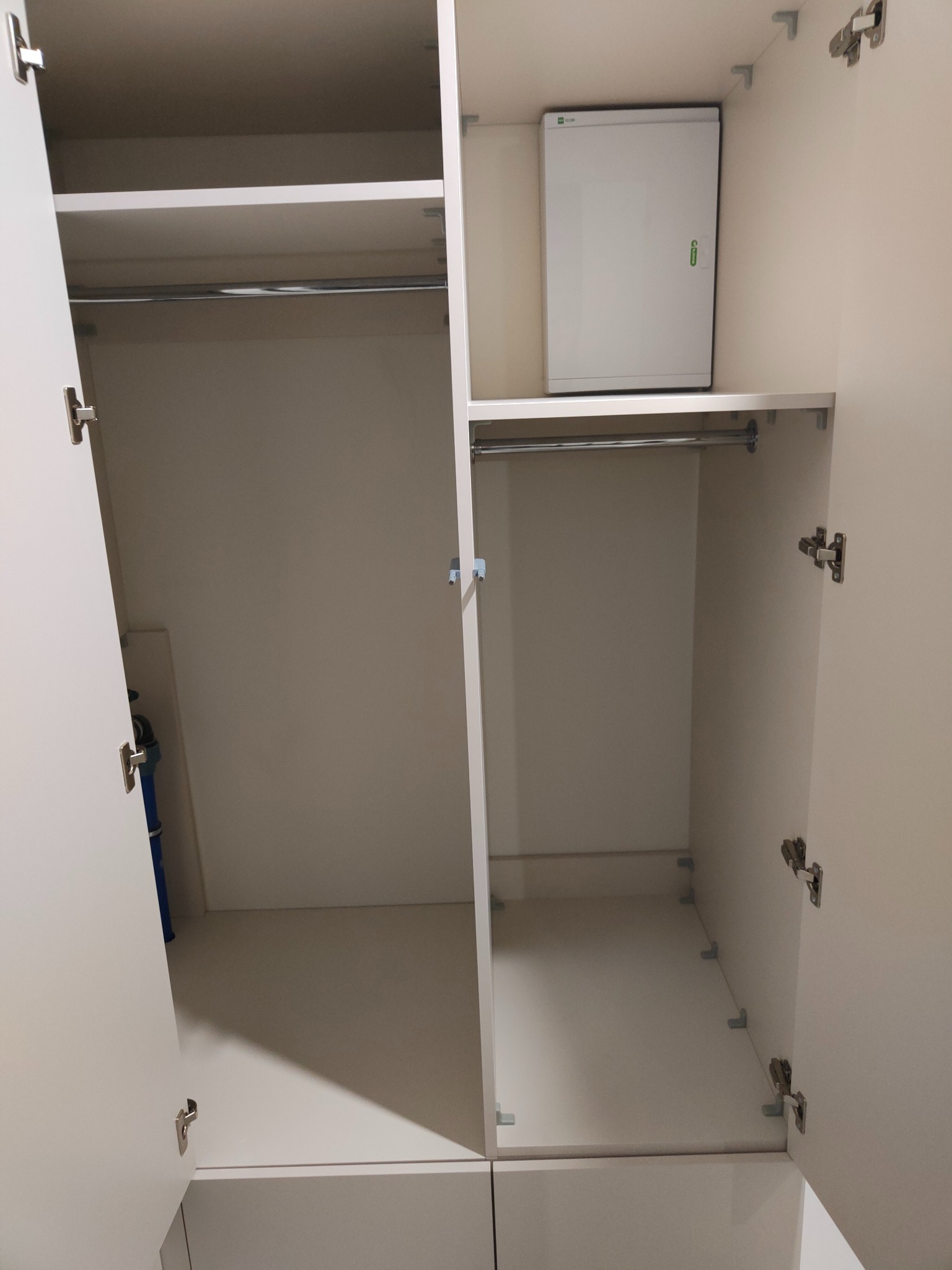 Wnętrze szafy wnękowej z drążkami na ubrania i szafką elektryczną, widok z otwartymi drzwiami, jasne wykończenie.