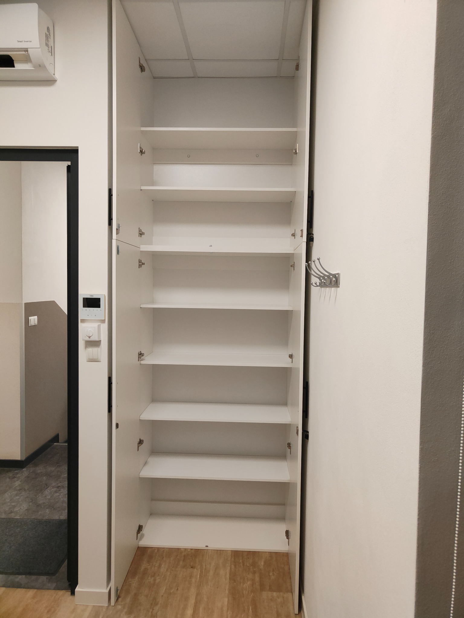 Biała szafa wnękowa z otwartymi drzwiami, widoczne półki. Na ścianie wieszak na ubrania i panel sterowania. Podłoga imitująca drewno. Minimalistyczny design.