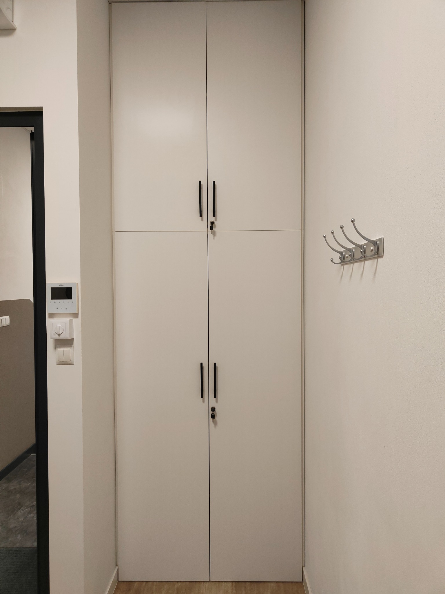 Biała szafa wnękowa z czarnymi, minimalistycznymi uchwytami, wkomponowana w jasne wnętrze przedpokoju. Obok wieszak na ubrania i panel sterowania domem.