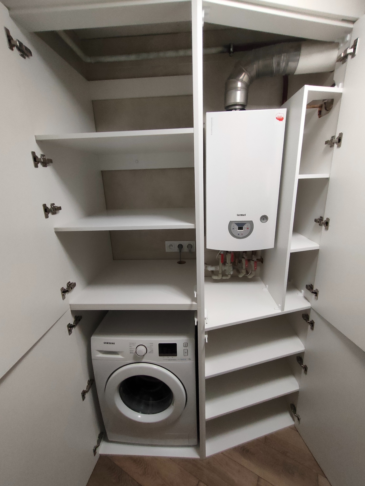 Biała szafa wnękowa z pralką i piecem gazowym. Widoczne półki i otwarte drzwi szafy. Minimalistyczny design, funkcjonalne zagospodarowanie przestrzeni.