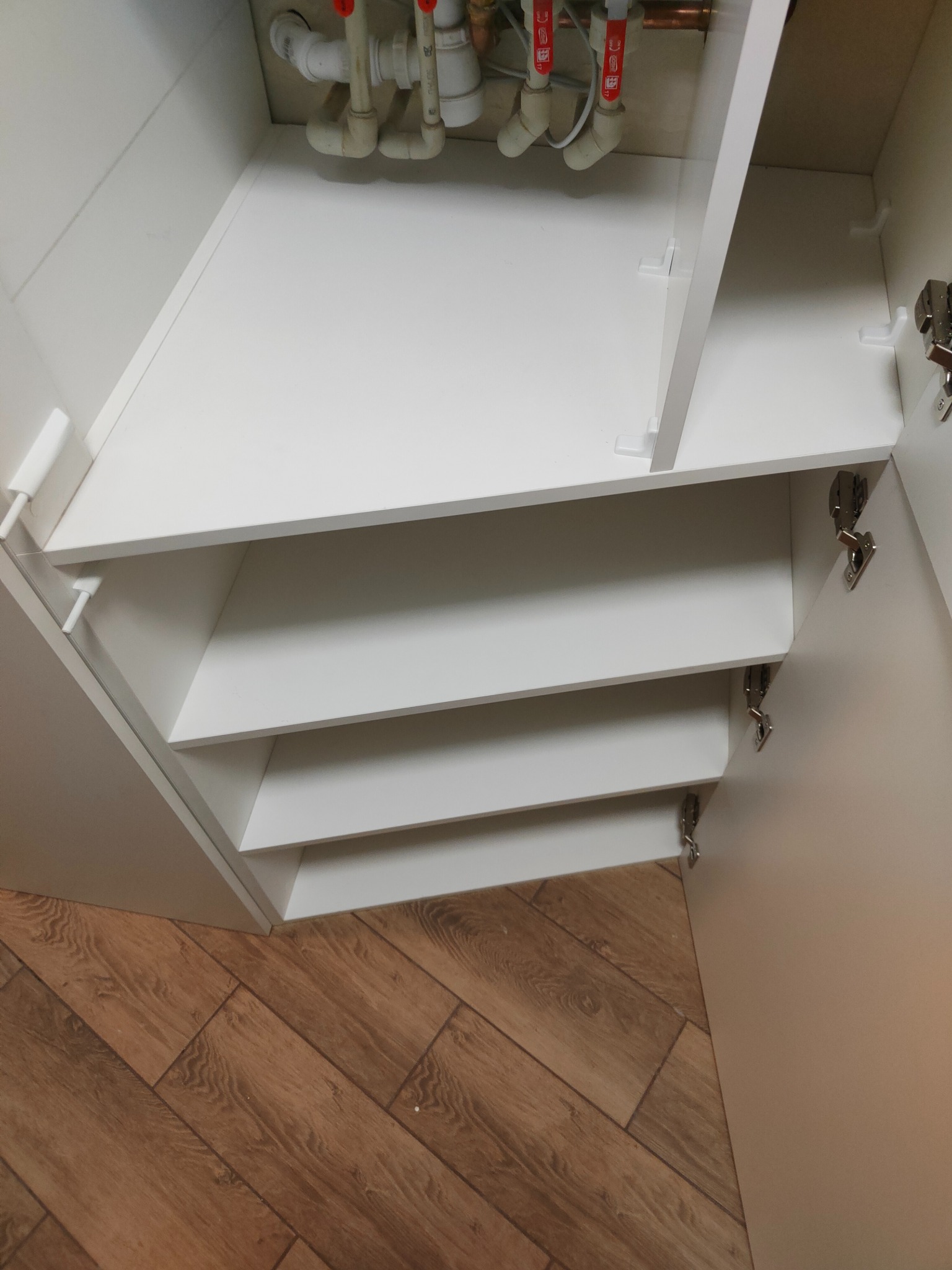 Wnętrze szafki z białymi półkami, widoczne rury instalacji wodnej u góry. Otwarta szafka z zawiasami, podłoga z paneli imitujących drewno. Ujęcie z góry.