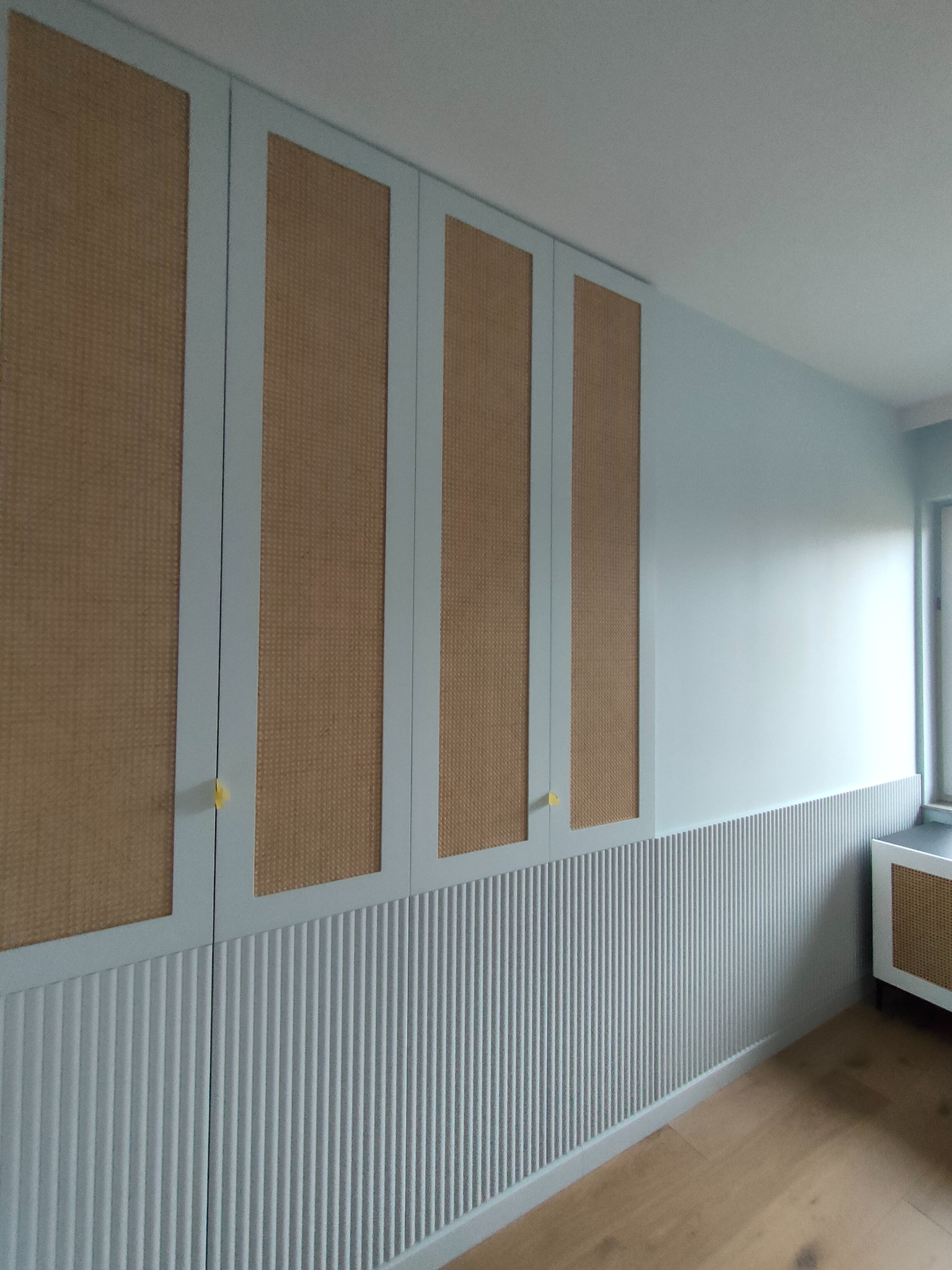 Błękitna szafa wnękowa z rattanowymi frontami i ryflowanym panelem ściennym, widok z dołu, podkreślający wysokość zabudowy i detale wykończenia.