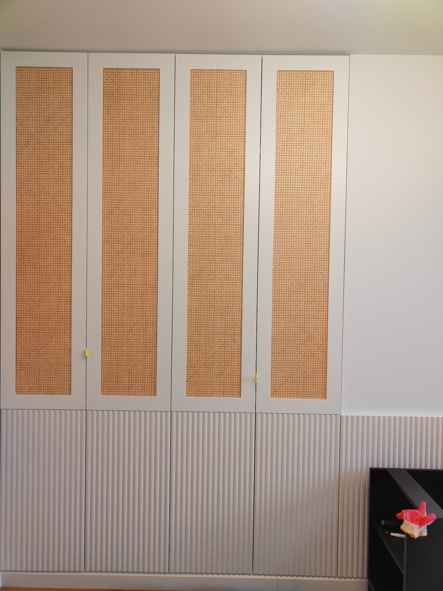 Biała szafa wnękowa z rattanowymi frontami i ryflowanym wykończeniem dolnej części. Minimalistyczny design, widoczne detale wykończenia i subtelne uchwyty.