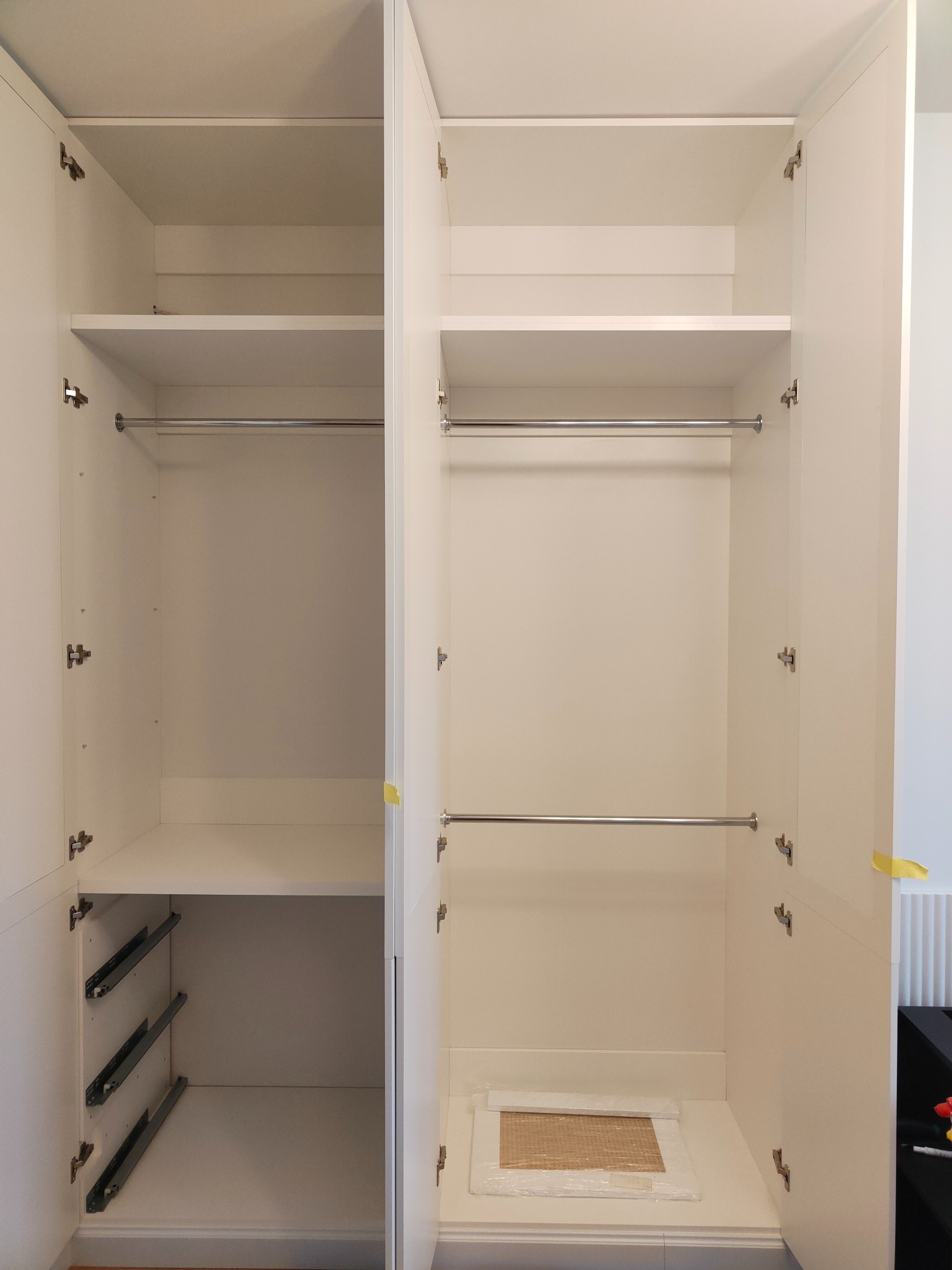 Wnętrze białej, otwartej szafy wnękowej z drążkami na ubrania i szufladami na prowadnicach. Widoczne zawiasy i elementy montażowe. Minimalistyczny design.