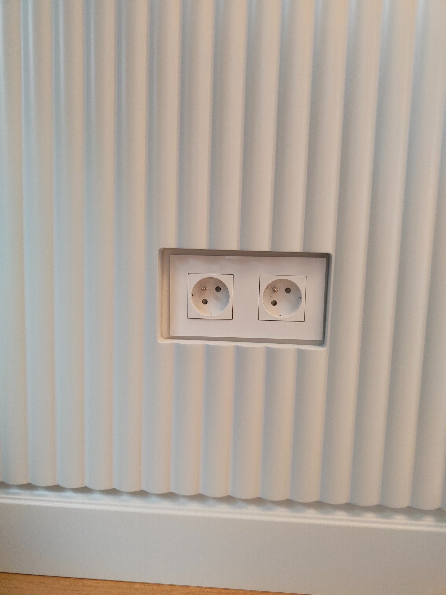 Biała, ryflowana ściana z dwoma gniazdkami elektrycznymi w białej ramce. Widoczna listwa przypodłogowa i fragment podłogi. Minimalistyczny design wnętrza.