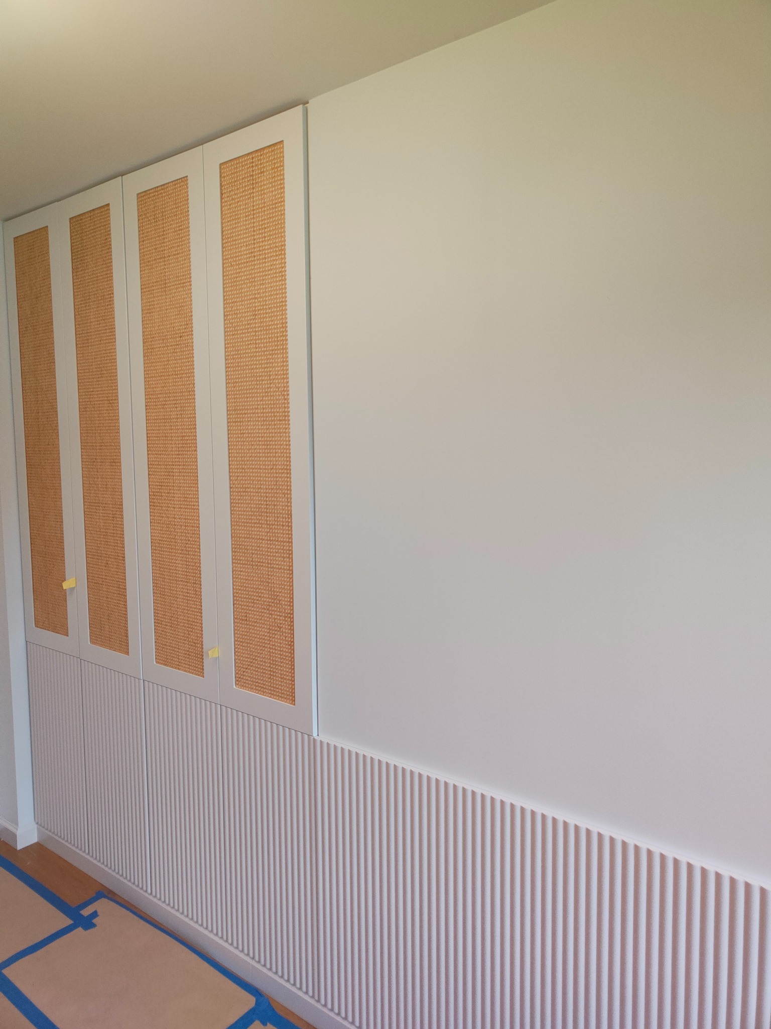 Biała szafa wnękowa z rattanowymi frontami i dekoracyjnymi listwami pionowymi, zabezpieczona taśmą malarską na czas prac wykończeniowych. Minimalistyczny design.