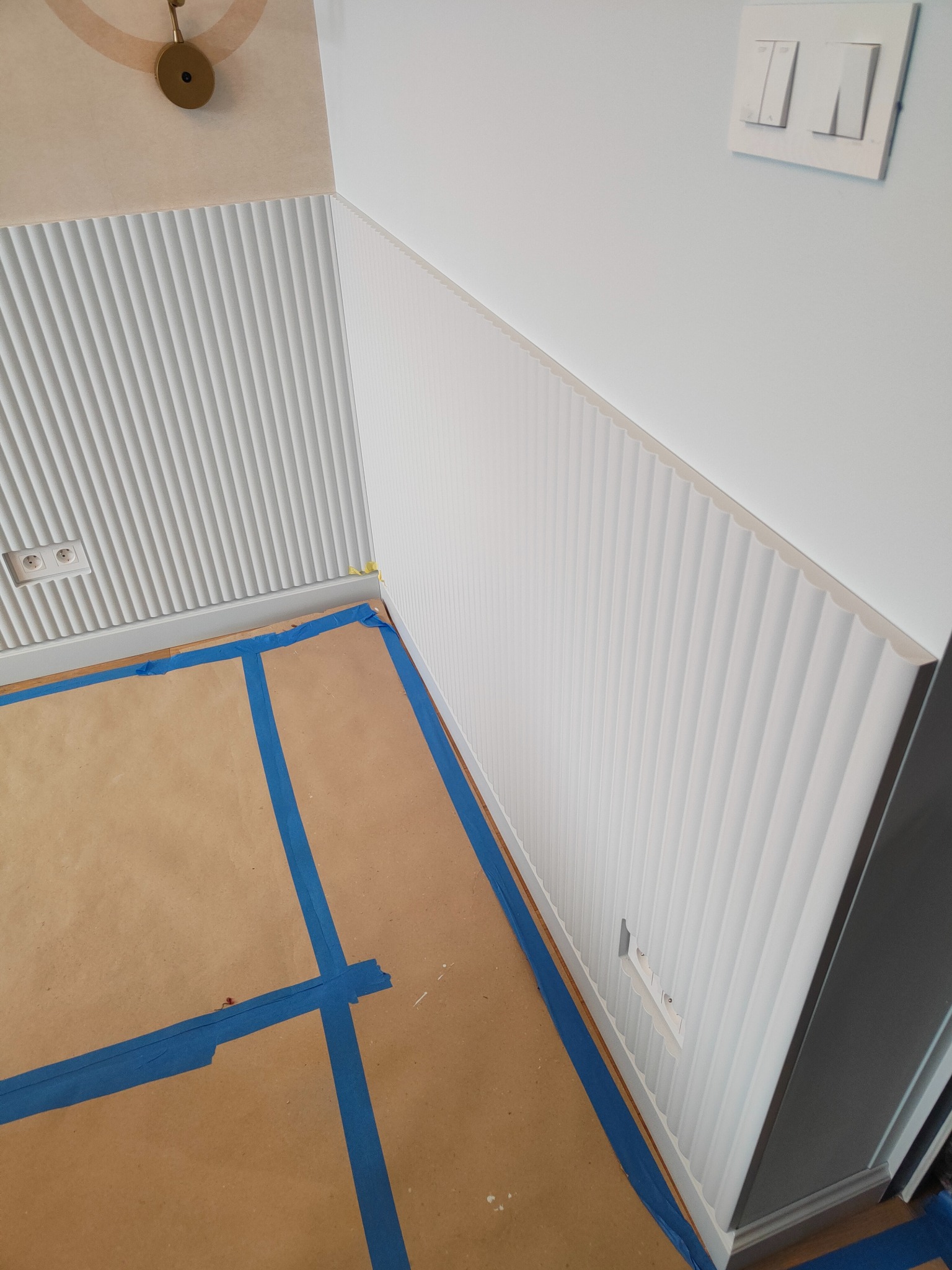 Montaż paneli ściennych z frezowaniem pionowym, zabezpieczona podłoga kartonem i taśmą malarską. Widoczne gniazdka elektryczne i włącznik światła.