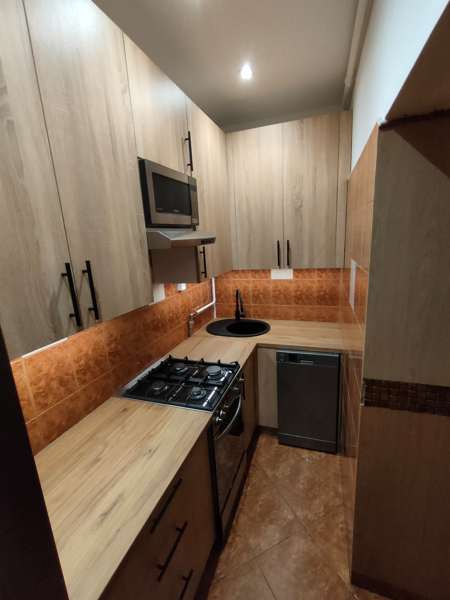 Nowoczesna kuchnia z drewnianymi frontami szafek, czarnymi uchwytami i brązowymi płytkami. Widoczna kuchenka gazowa i zlew. Ujęcie z perspektywy.