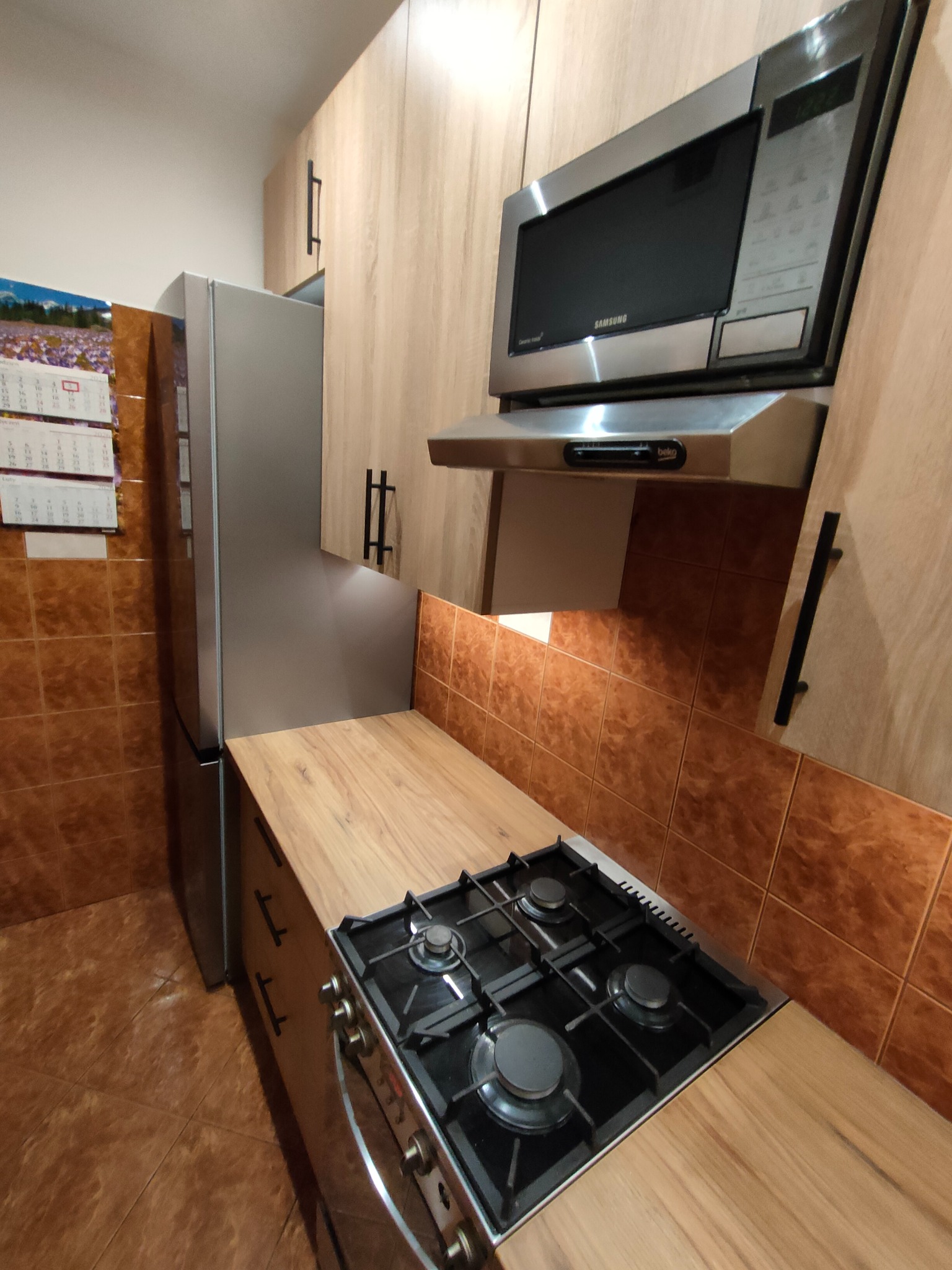 Nowoczesna kuchnia z drewnianymi frontami, srebrną lodówką i kuchenką gazową. Uwagę przykuwa brązowa glazura na ścianie i kalendarz na lodówce.