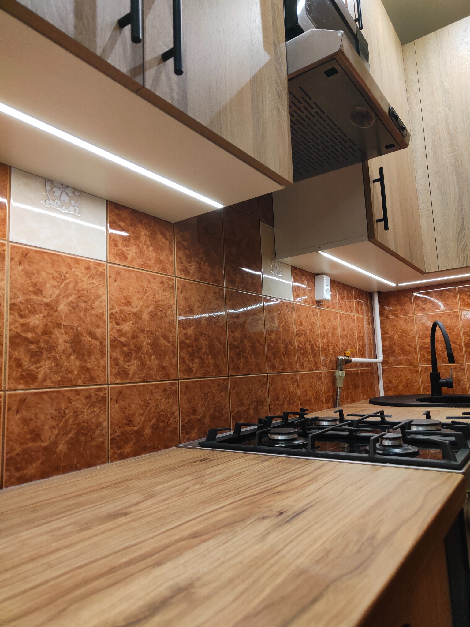 Nowoczesna kuchnia z drewnianym blatem, brązowymi płytkami i oświetleniem LED pod szafkami. Widoczna płyta gazowa i czarna bateria. Ujęcie z perspektywy blatu.