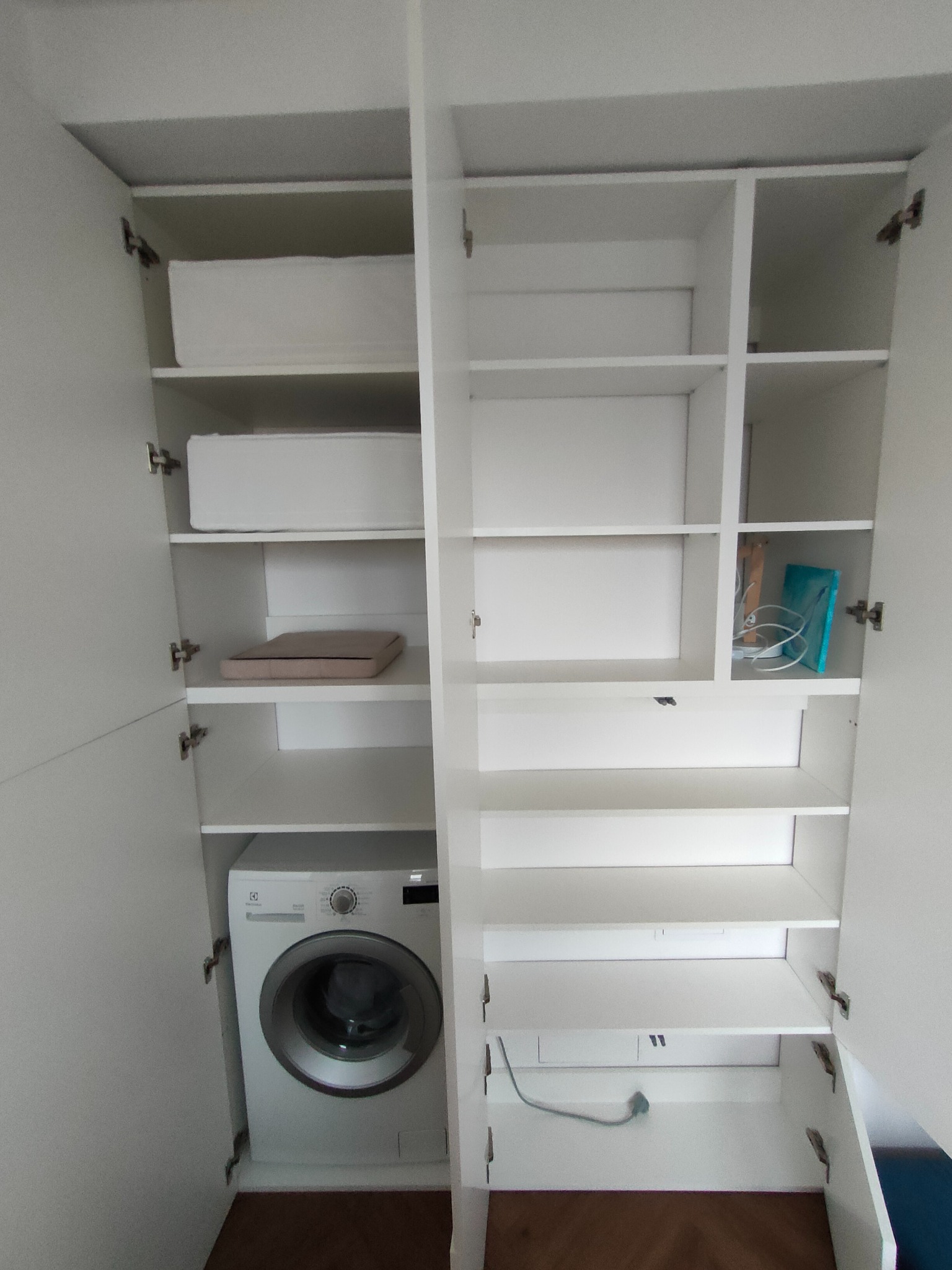Biała szafa wnękowa z pralką i półkami, otwarte drzwi ukazują wnętrze. Widoczne akcesoria i instalacja elektryczna. Minimalistyczny design, jasne oświetlenie.