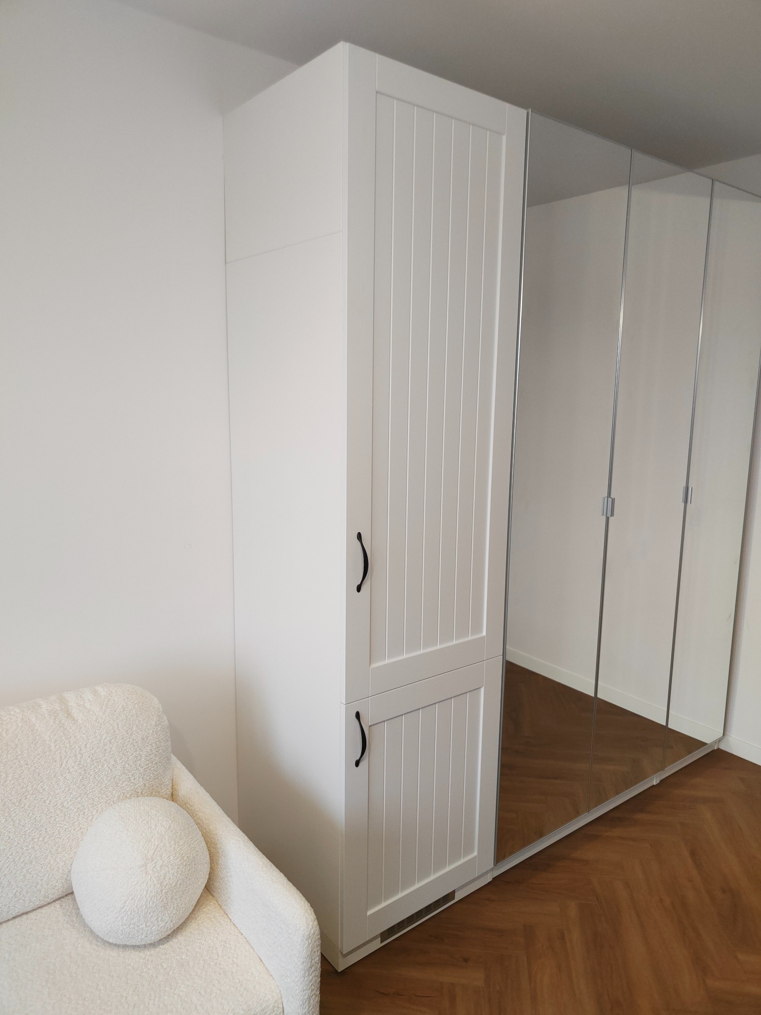 Biała szafa wnękowa z lustrzanymi drzwiami i frezowanymi frontami, uzupełniona kremowym fotelem z okrągłą poduszką na tle parkietu ułożonego w jodełkę.