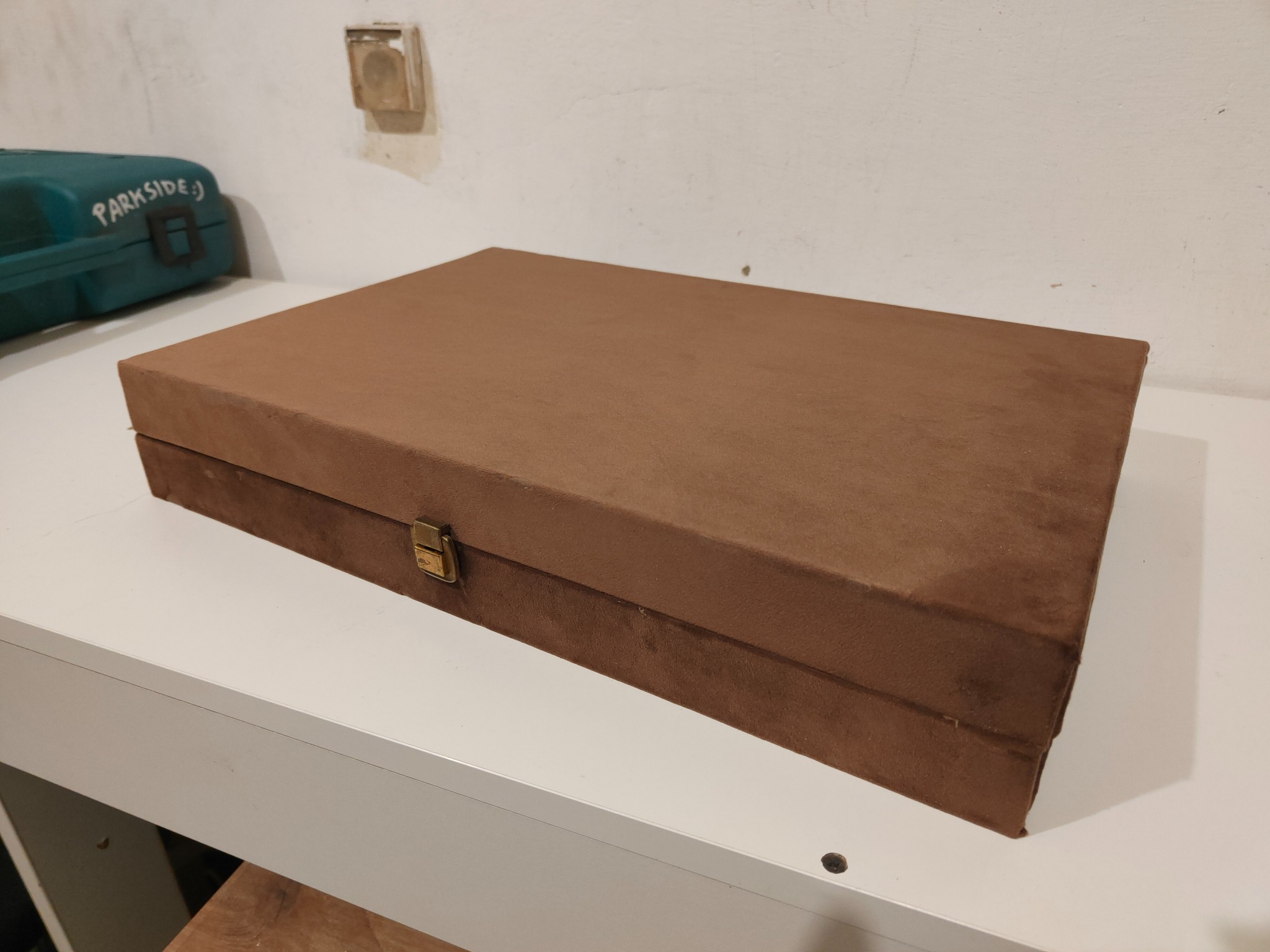 Brązowe, prostokątne pudełko z metalowym zapięciem na białym blacie. W tle ściana z widocznym gniazdkiem i zielona skrzynka narzędziowa Parkside.