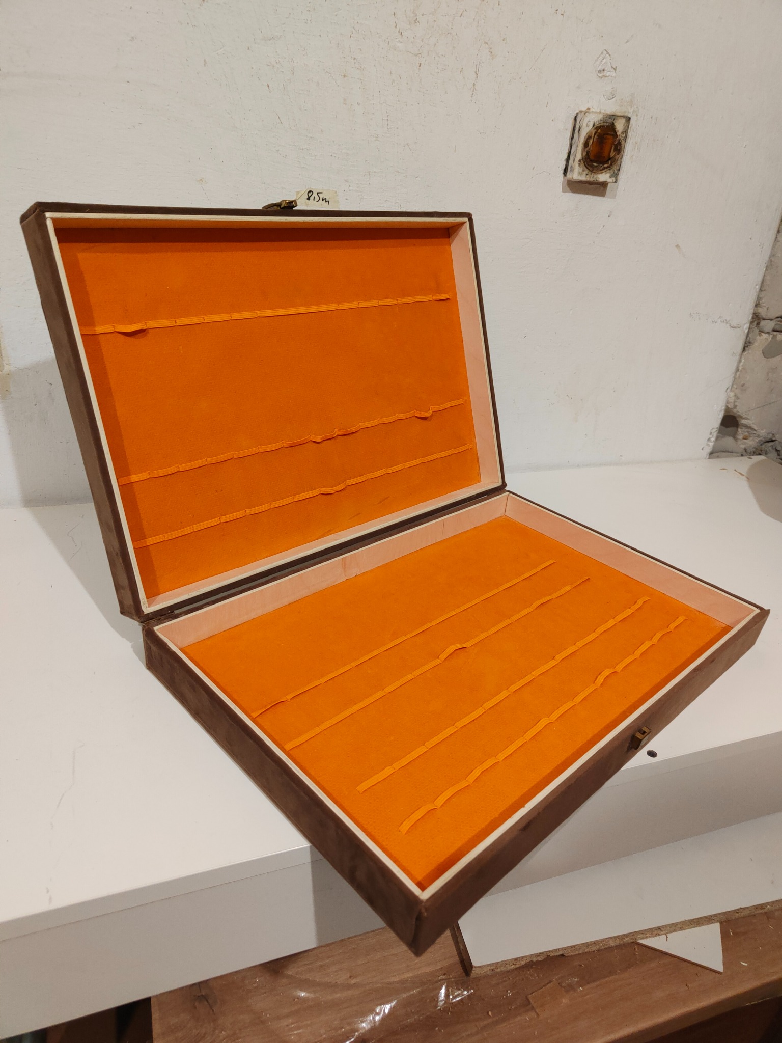 Otwarta, brązowa szkatułka z pomarańczowym wnętrzem, widoczne przegródki. Szkatułka stoi na białym blacie, w tle ściana z widocznymi niedoskonałościami.