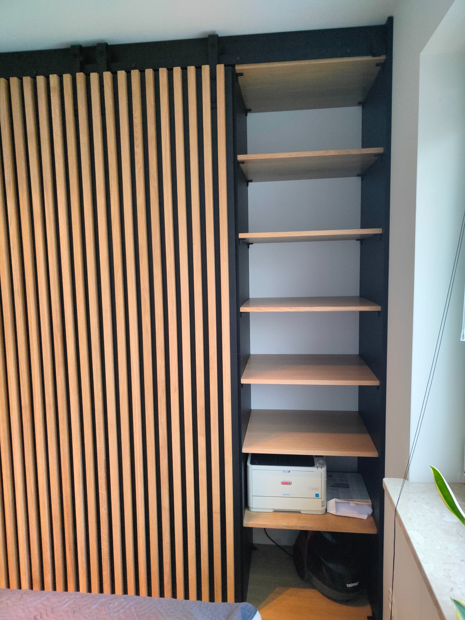 Szafa wnękowa z przesuwnymi drzwiami lamelowymi z drewna, czarna rama, półki z jasnego drewna. Na półkach drukarka i torba. Nowoczesny design.