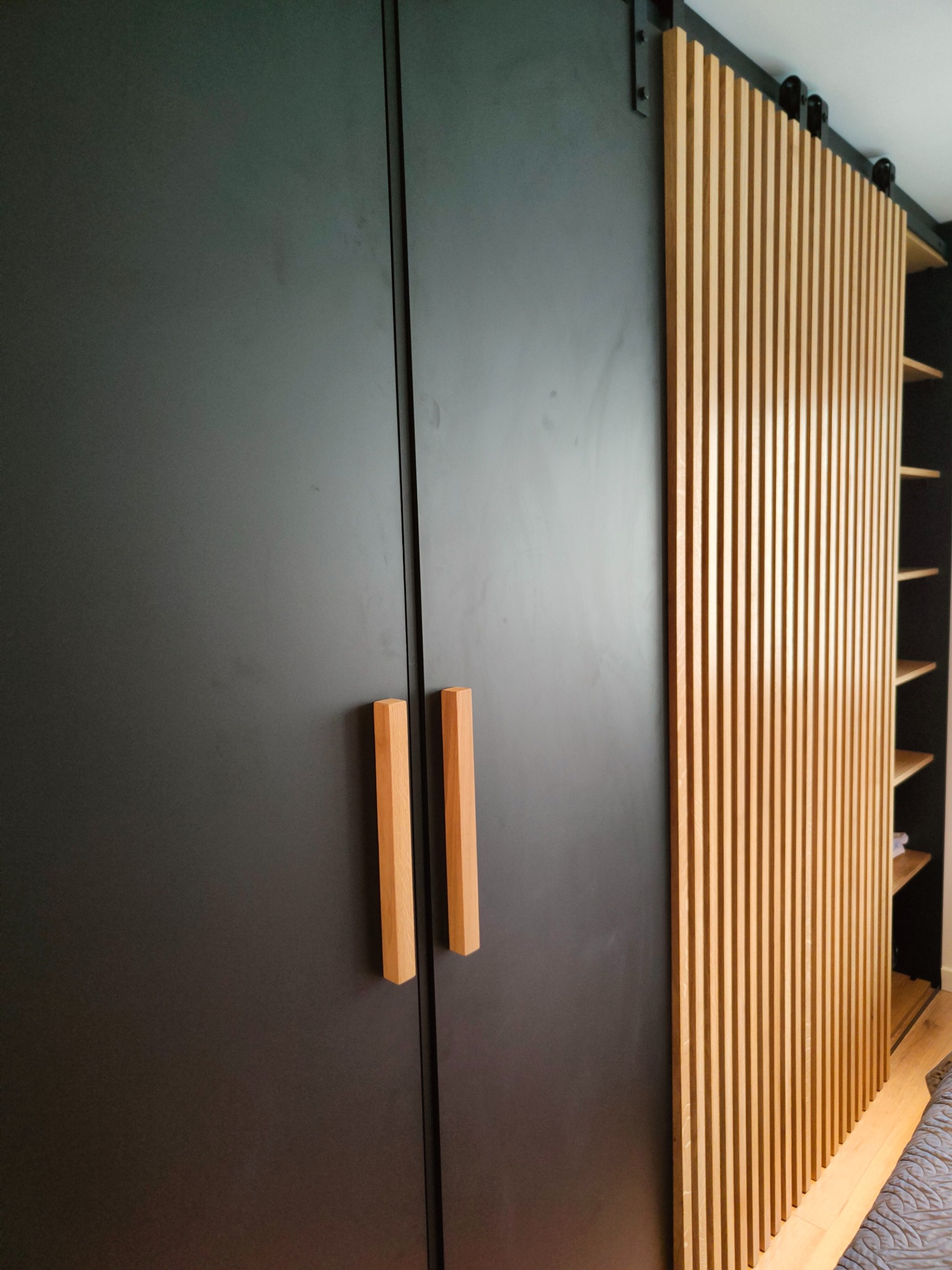 Nowoczesna szafa wnękowa z przesuwnymi drzwiami lamelowymi i drewnianymi uchwytami, w minimalistycznym wnętrzu. Czarny mat frontów kontrastuje z jasnym drewnem.