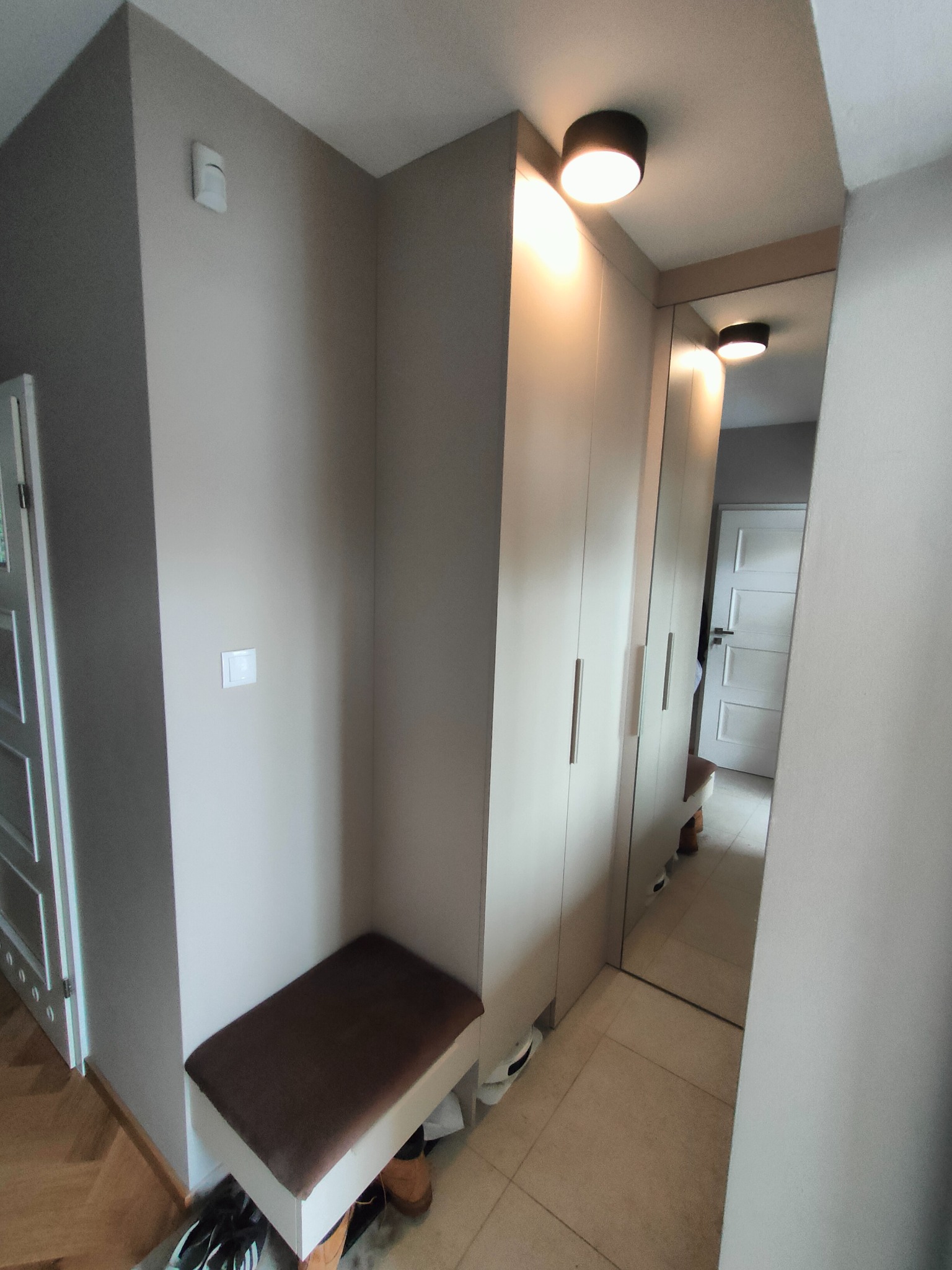 Jasny przedpokój z szafą wnękową z lustrzanymi drzwiami, oświetleniem punktowym i siedziskiem z brązowym obiciem. Widoczne buty pod siedziskiem.