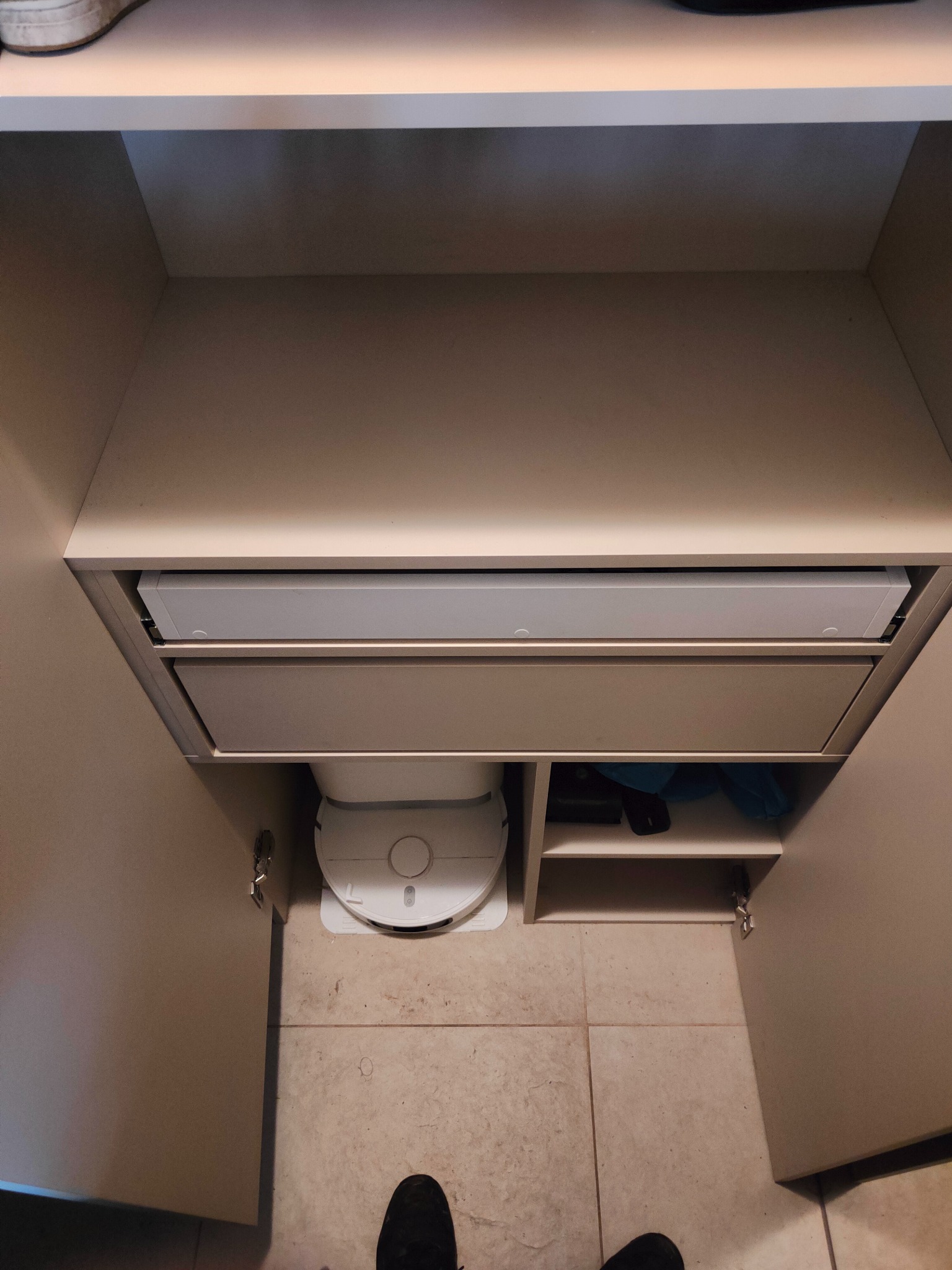 Wnętrze szafy wnękowej z szufladami i półkami, na podłodze robot sprzątający. Widoczne zawiasy drzwi i fragment buta osoby robiącej zdjęcie.