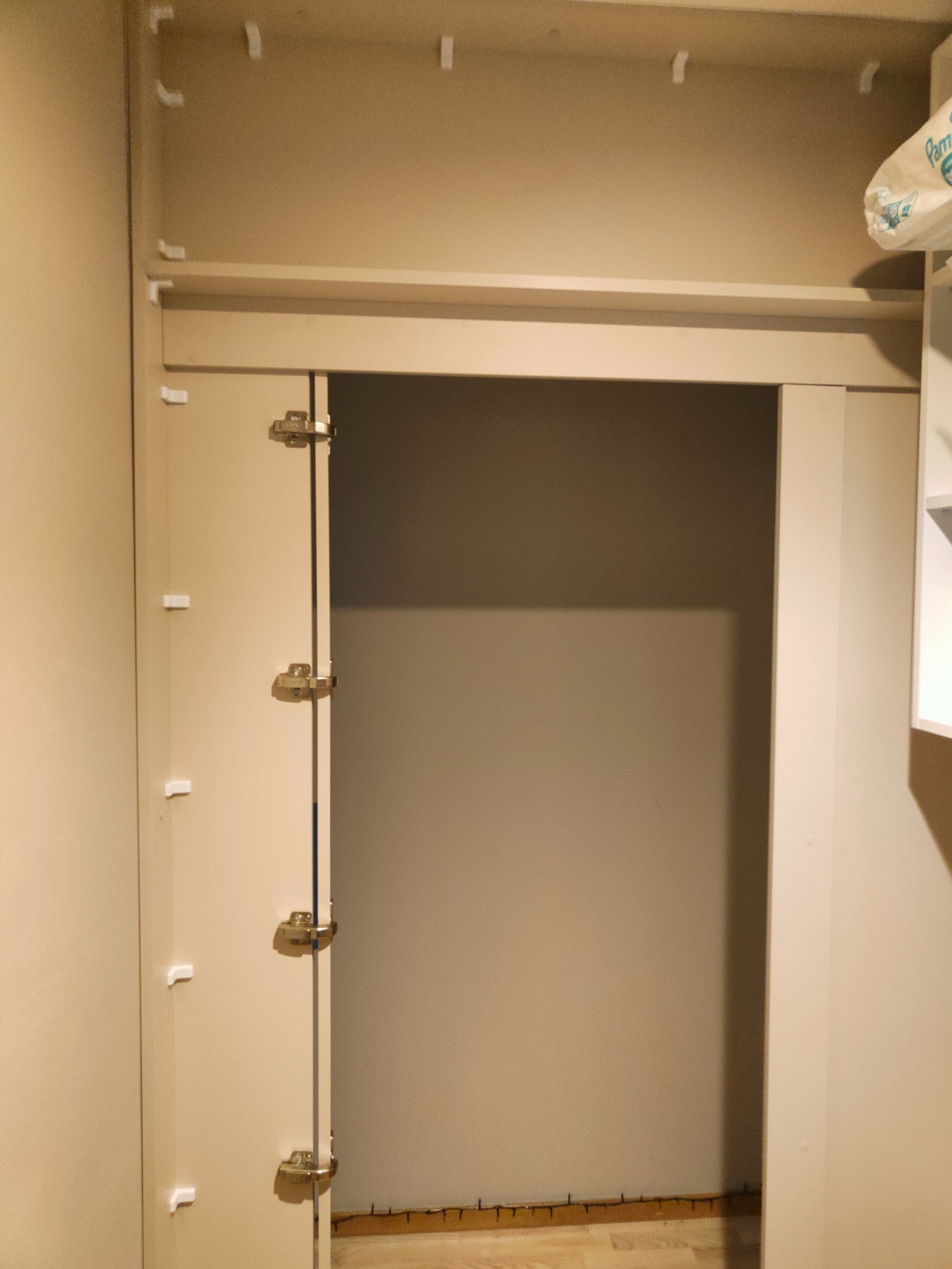 Otwarta szafa wnękowa z widocznymi zawiasami i elementami mocującymi, ukazująca puste wnętrze z drewnianą podłogą. Minimalistyczny design, beżowe wykończenie.