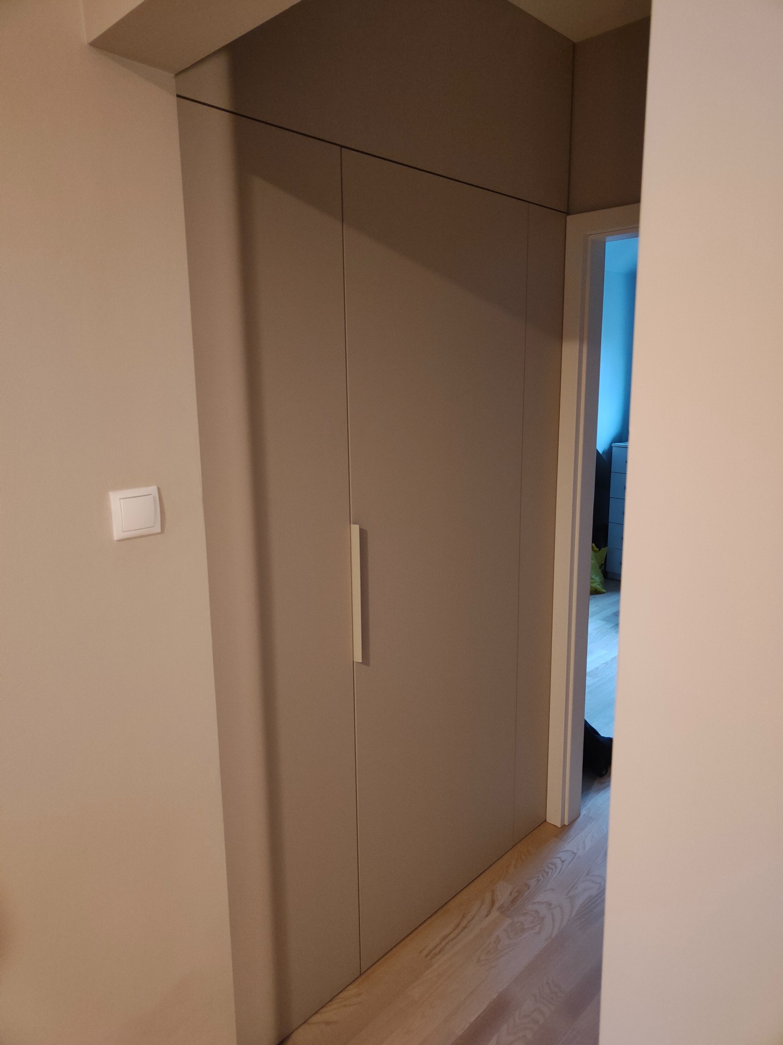 Minimalistyczna szafa wnękowa w odcieniach beżu, z długim, pionowym uchwytem. Widoczny fragment parkietu i wnętrze pokoju przez otwarte drzwi.