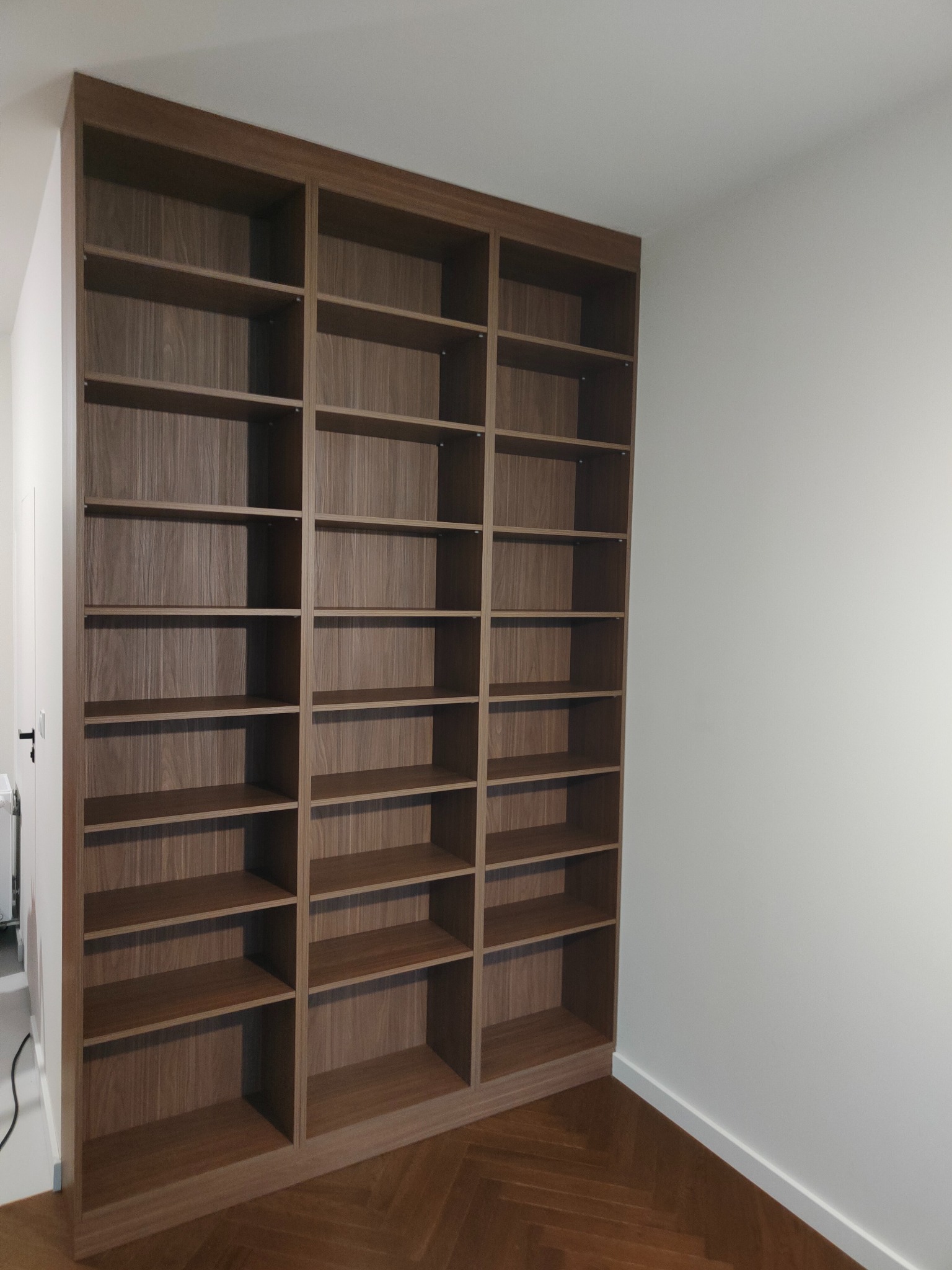 Wysoka, drewniana biblioteczka na wymiar z licznymi półkami, wkomponowana we wnękę, z parkietem ułożonym w jodełkę na podłodze. Minimalistyczne wnętrze.